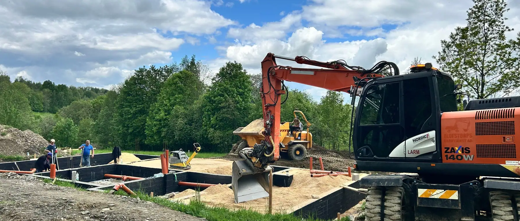 Koparka kołowa Hitachi Zaxis 140W z tiltrotatorem przy szalunkach fundamentu, obok wozidło Wacker Neuson i pracownicy na budowie