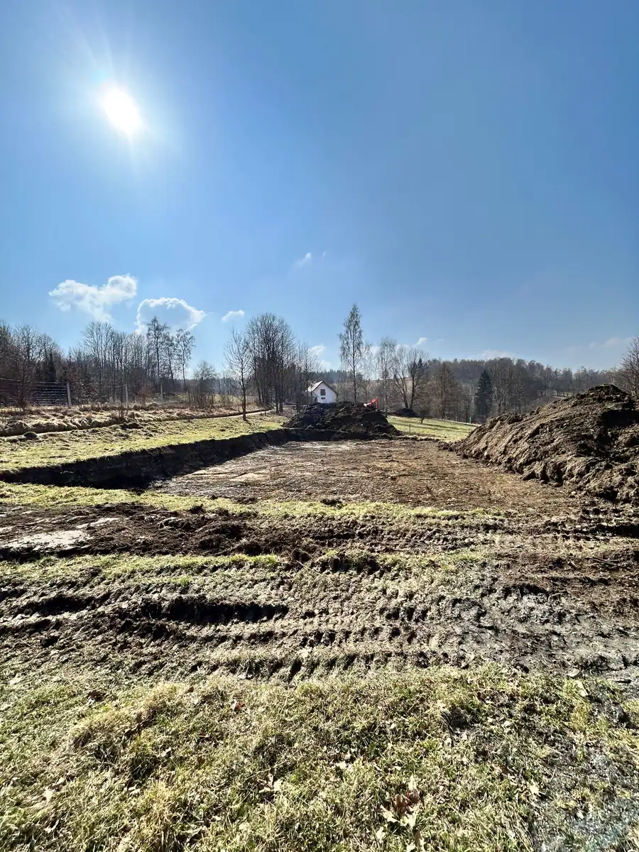 Szeroki wykop pod fundament na działce ze spadkiem, pryzma humusu i koparka kołowa w tle