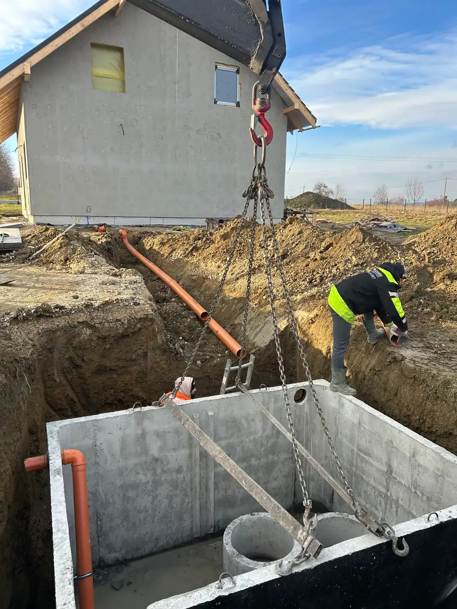 Betonowy zbiornik na szambo opuszczany żurawiem do wykopu przy nowym domu, obok rura kanalizacyjna