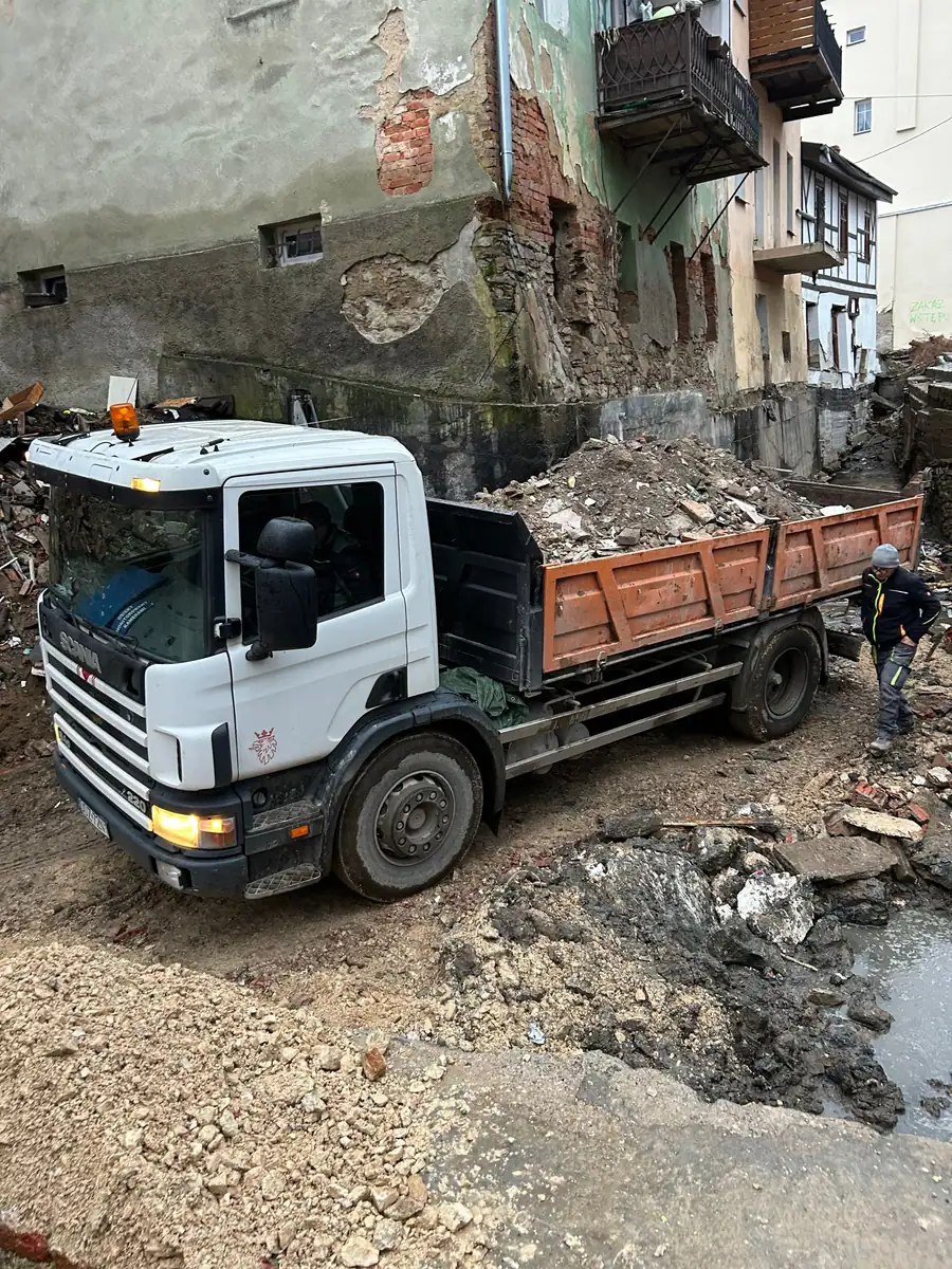 Wywrotka Scania 220 4×2 z gruzem w wąskiej uliczce między uszkodzonymi kamienicami